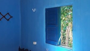 Una habitación con paredes azules, una cama individual con colcha a cuadros, una ventana con contraventanas azules y una mesita con una vela.