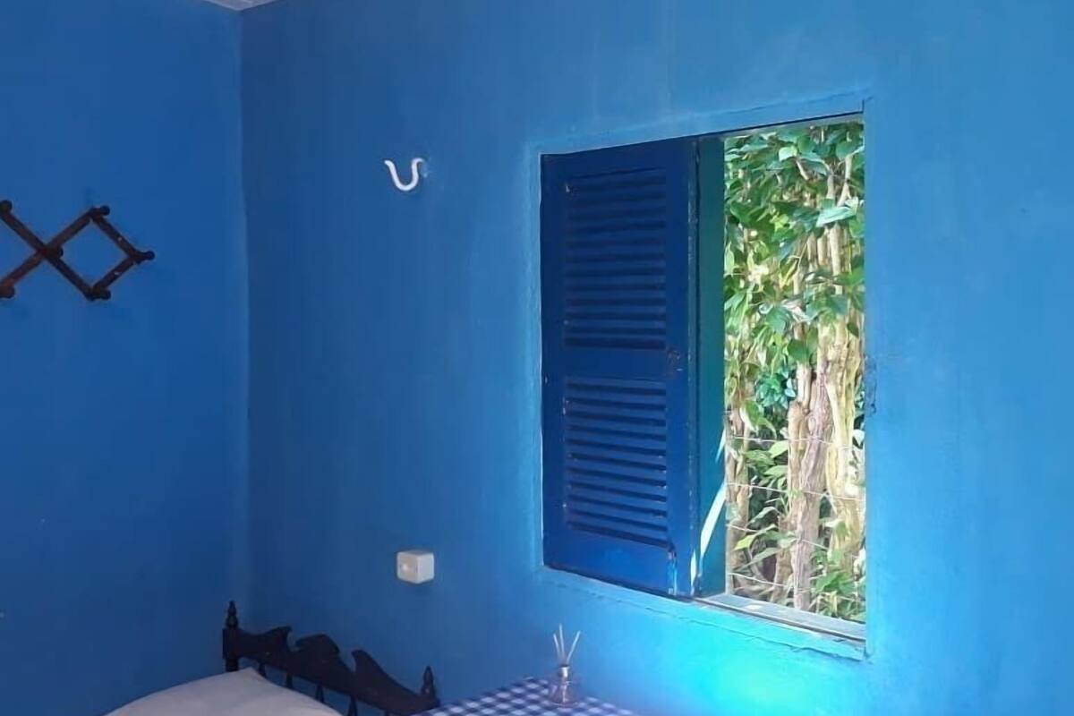 Ein Zimmer mit blauen Wänden, einem Einzelbett mit karierter Bettdecke, einem Fenster mit blauen Rollläden und einem kleinen Tisch mit einer Kerze.