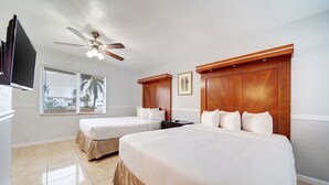 Quarto de hotel com duas camas, ventoinha no teto, televisão e janela com persianas.