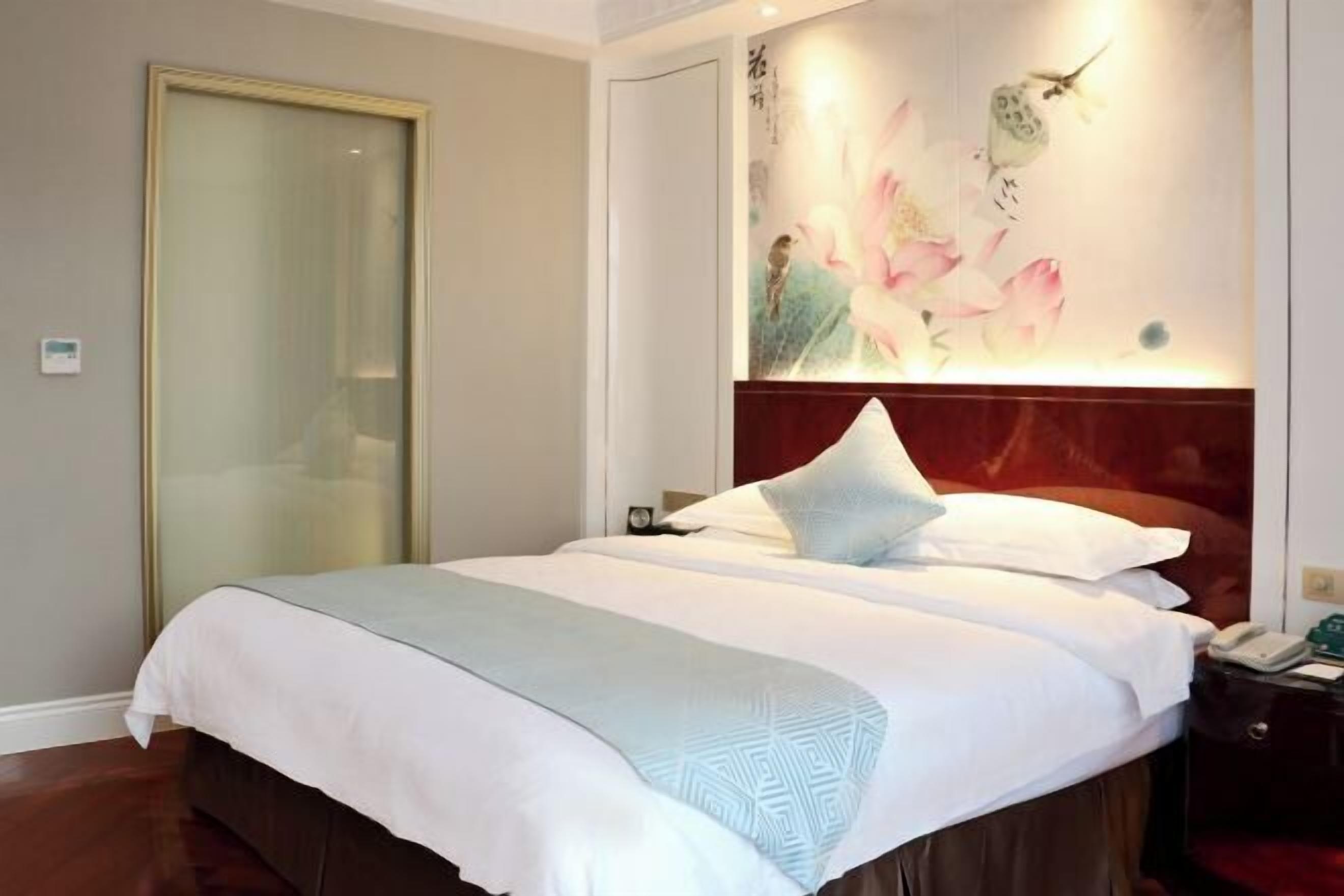 A hotel room with a large bed, a wooden headboard, a painting of flowers on the wall, and a sliding door.