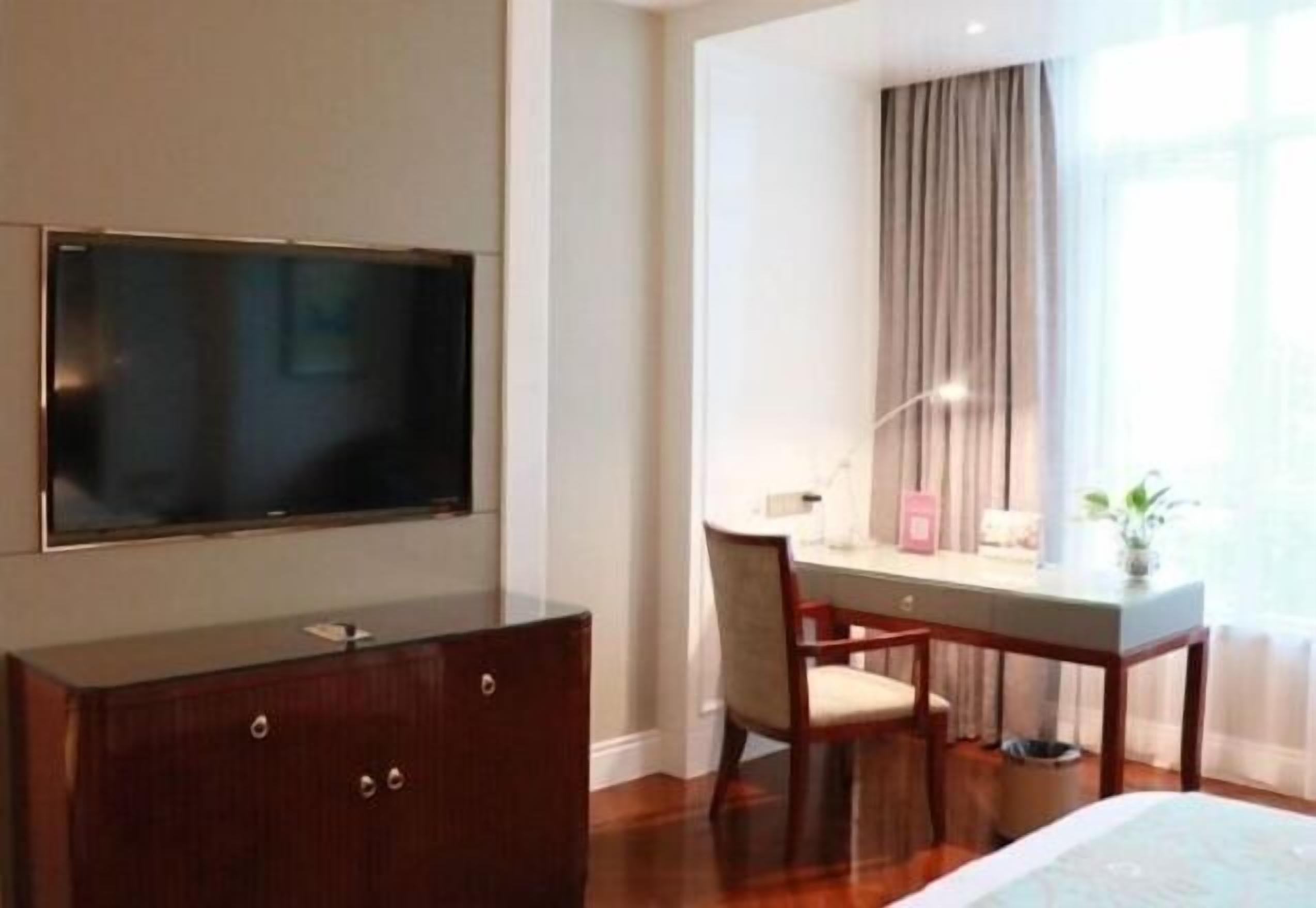 Ein Hotelzimmer mit Flachbildfernseher an der Wand, einem Holztisch mit Stuhl und einem Bett mit weißer Bettdecke.