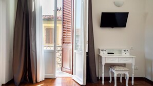 A room with a wooden floor, a white desk with drawers, a television mounted on the wall, and a door leading to an outdoor area with a balcony. - Live Florence Guesthouse (Florence)