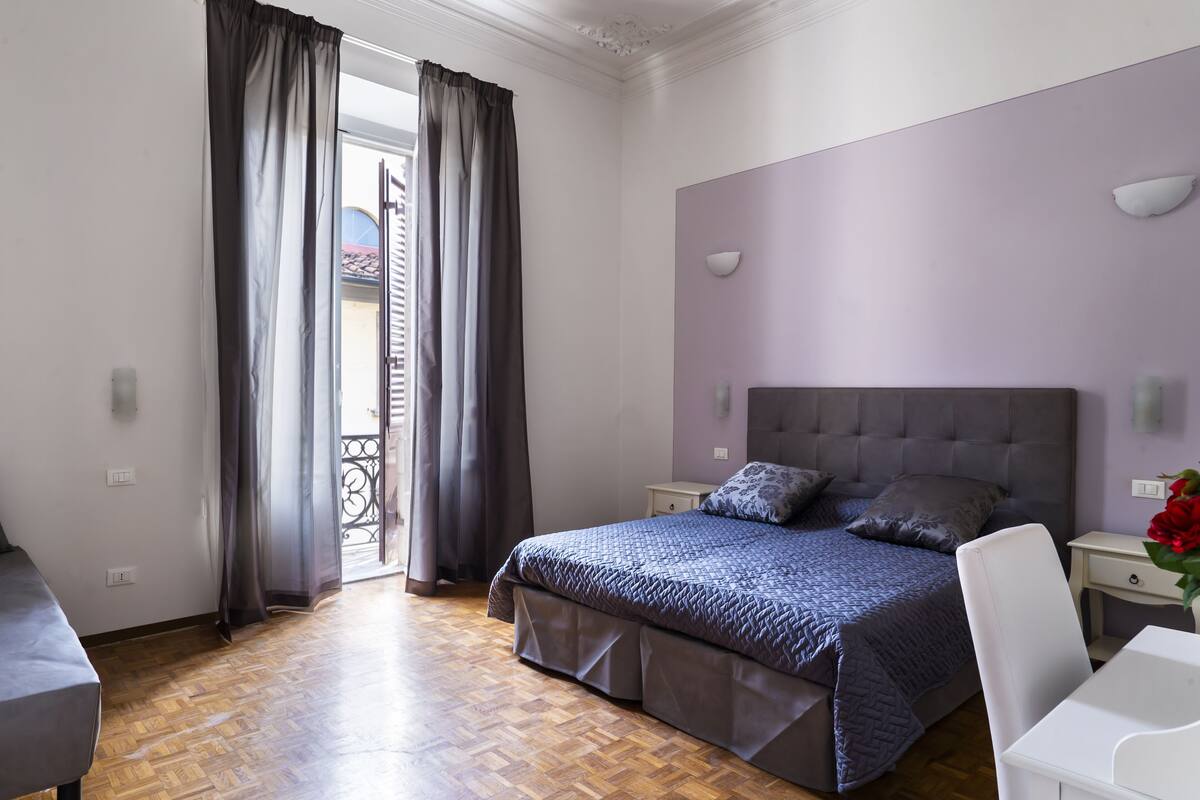 Een slaapkamer met een bed, nachtkastjes, een stoel en uitzicht door een raam met gordijnen.