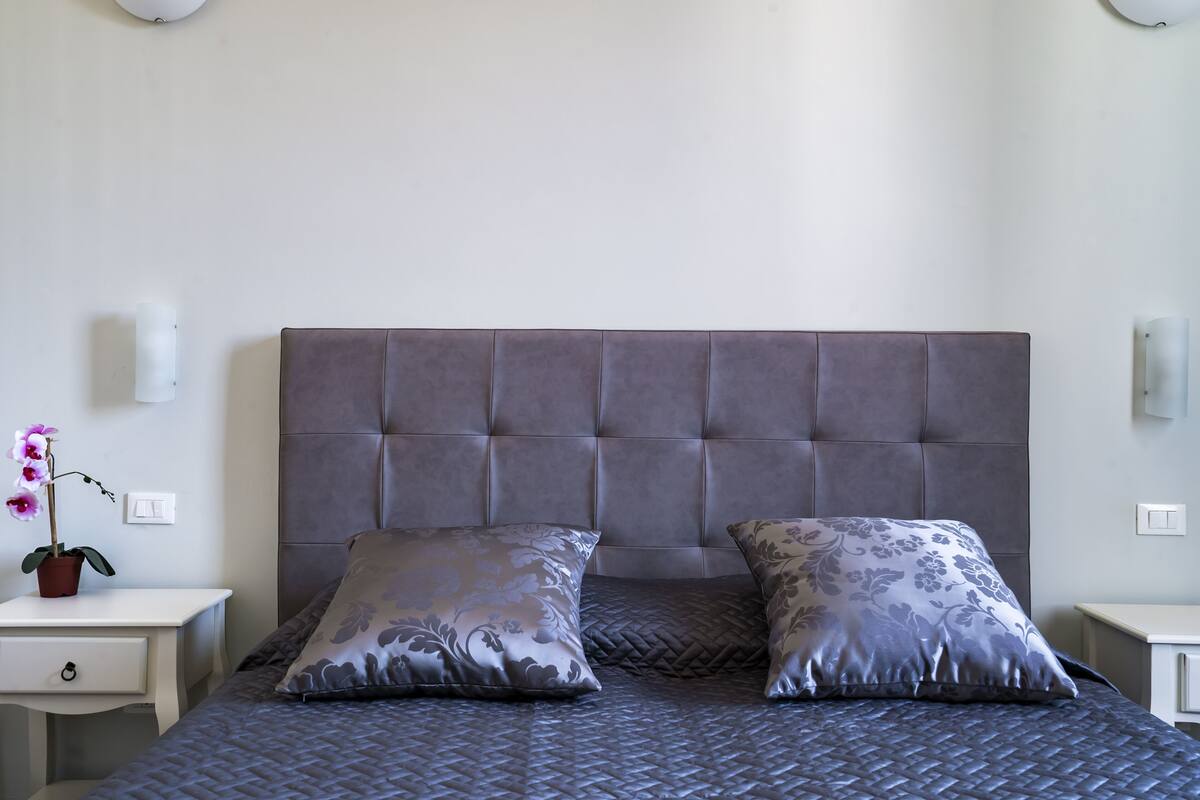 Een slaapkamer met een gebobbelde hoofdbord, twee kussens met een patroon, een donkere gewatteerde sprei, een nachtkastje met een potorkidee en een witte lade.