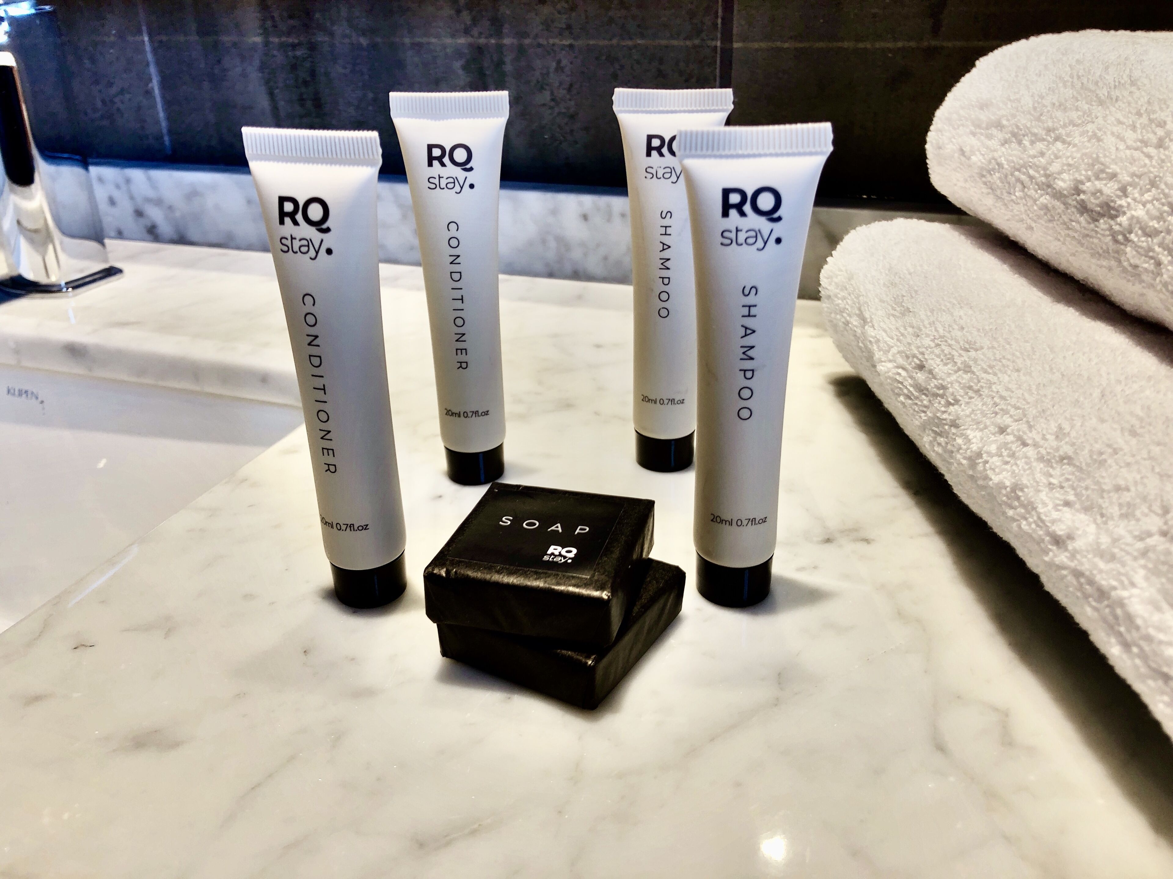 Cuatro tubos de RQ stay, acondicionador, champú y jabón sobre una superficie de mármol, con un estuche negro para jabón y una toalla blanca al fondo.