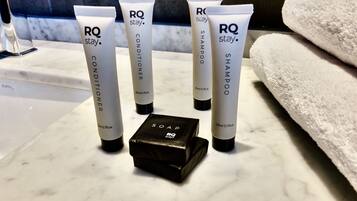 Cuatro tubos de RQ stay, acondicionador, champú y jabón sobre una superficie de mármol, con un estuche negro para jabón y una toalla blanca al fondo.