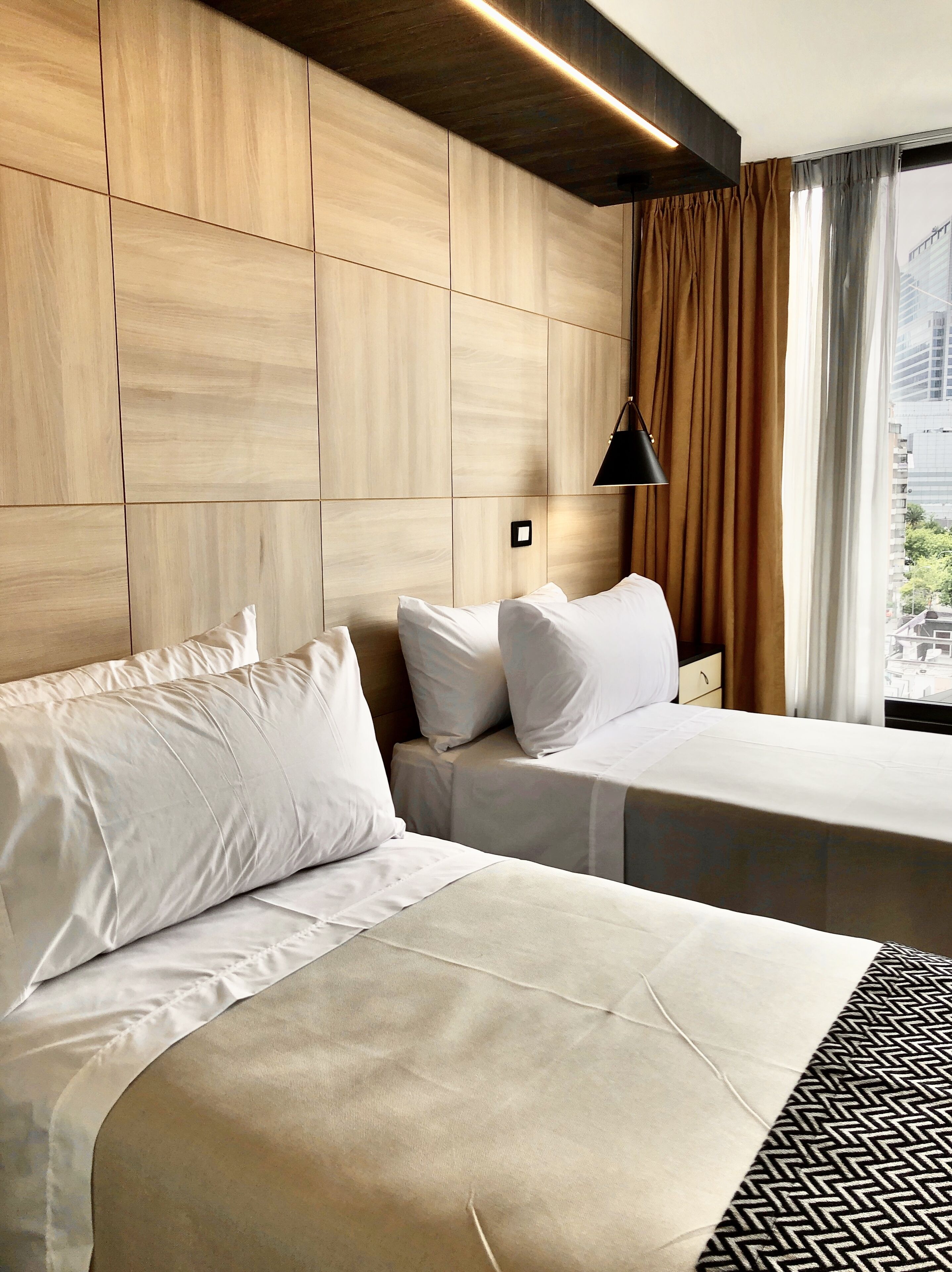 Habitación de hotel con dos camas, paredes de paneles de madera, una lámpara colgante negra y vistas a edificios a través del ventanal.