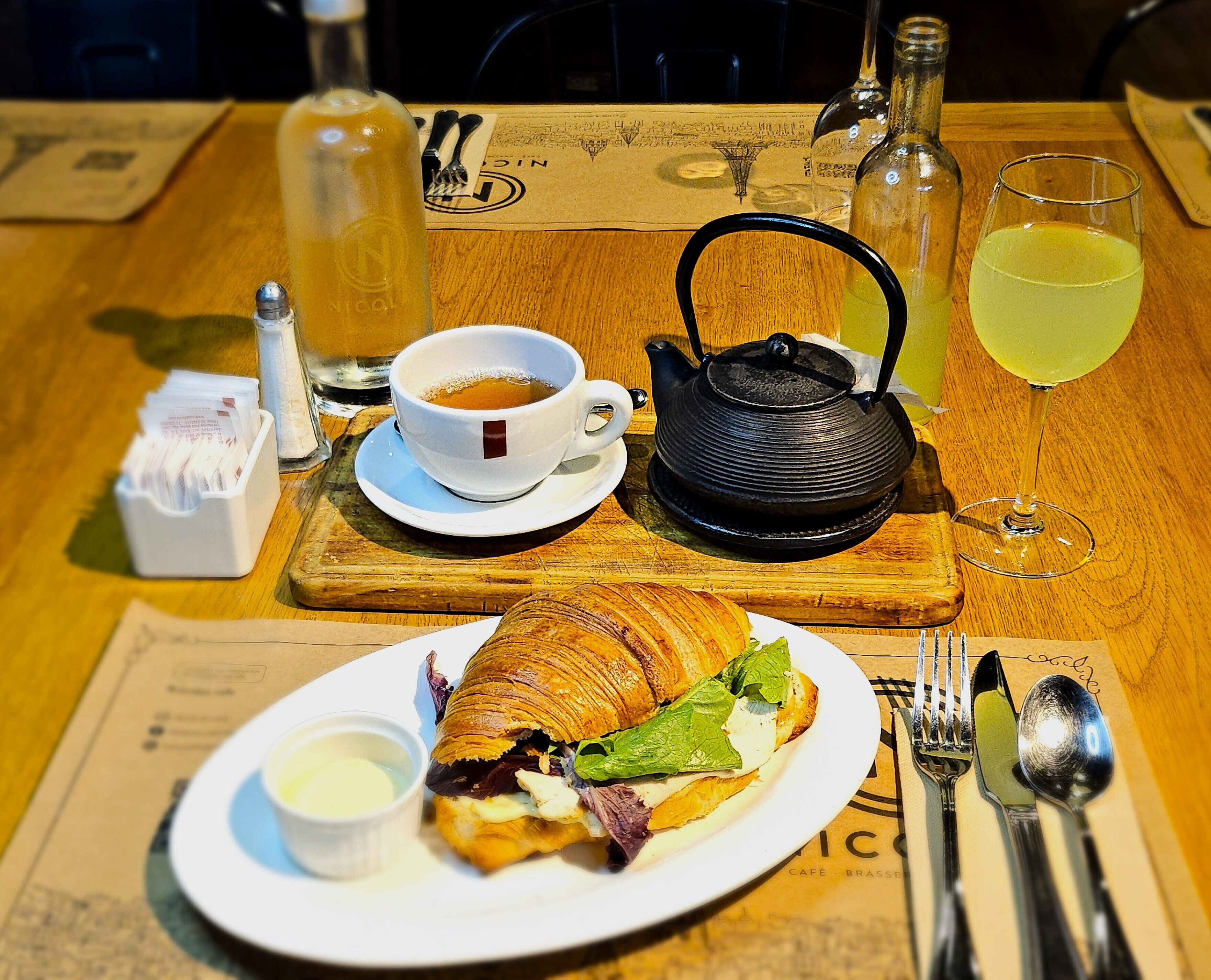 A table with a croissant sandwich, a cup of tea, a teapot, a bottle, and a glass of juice.