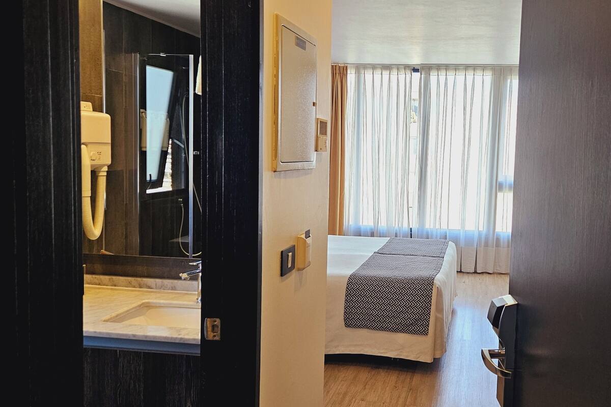 A hotel room with a bed, a bathroom with a mirror and sink, and a window with curtains.