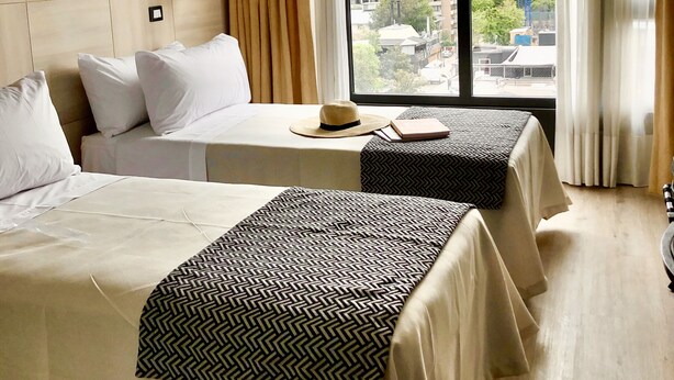 A hotel room with two beds, a large window with curtains, a city view, and a modern design. - RQ Santa Magdalena (Santiago)