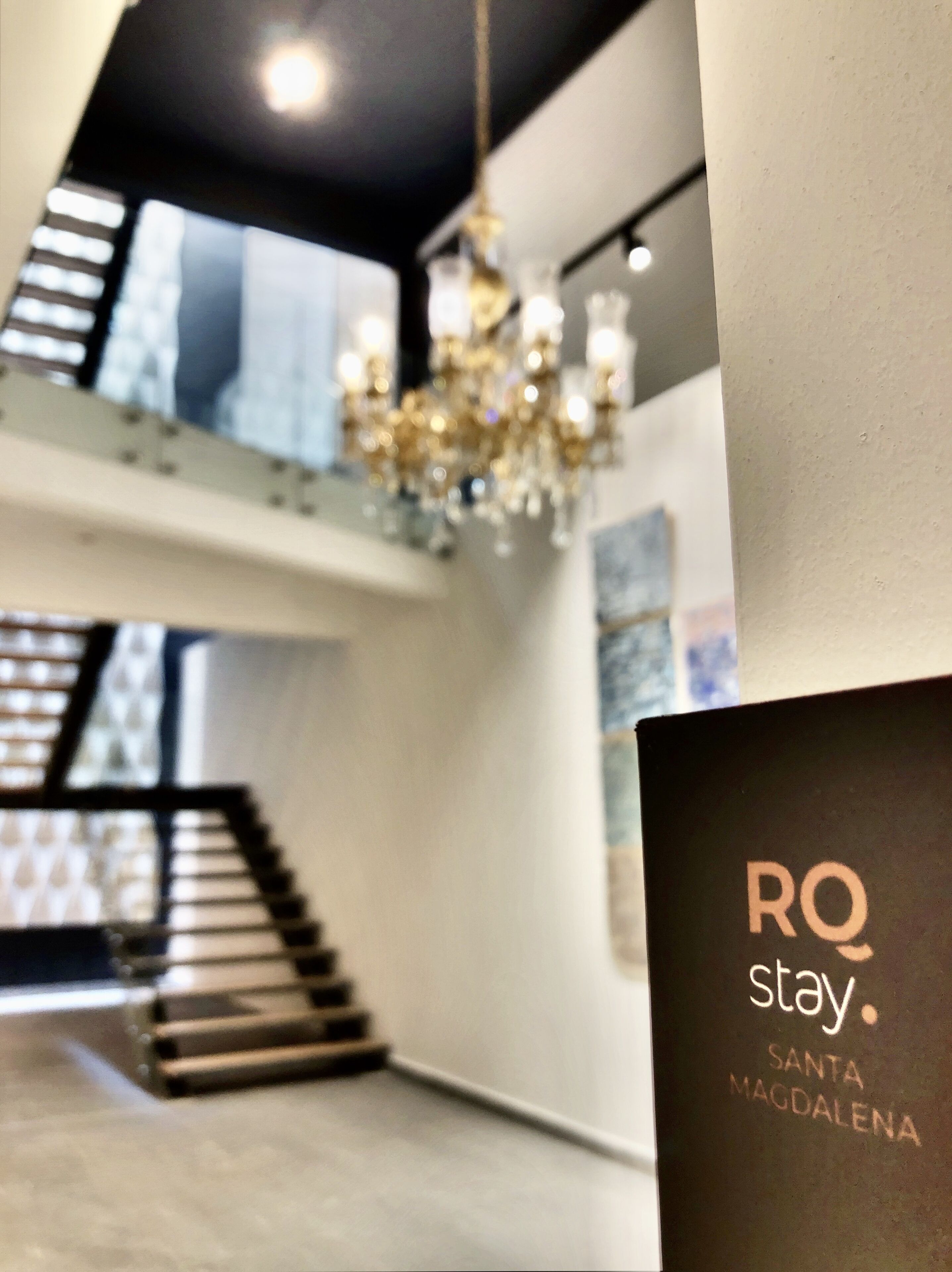 A modern interior with a staircase, chandelier, and a sign reading "RQ stay Santa Magdalena".