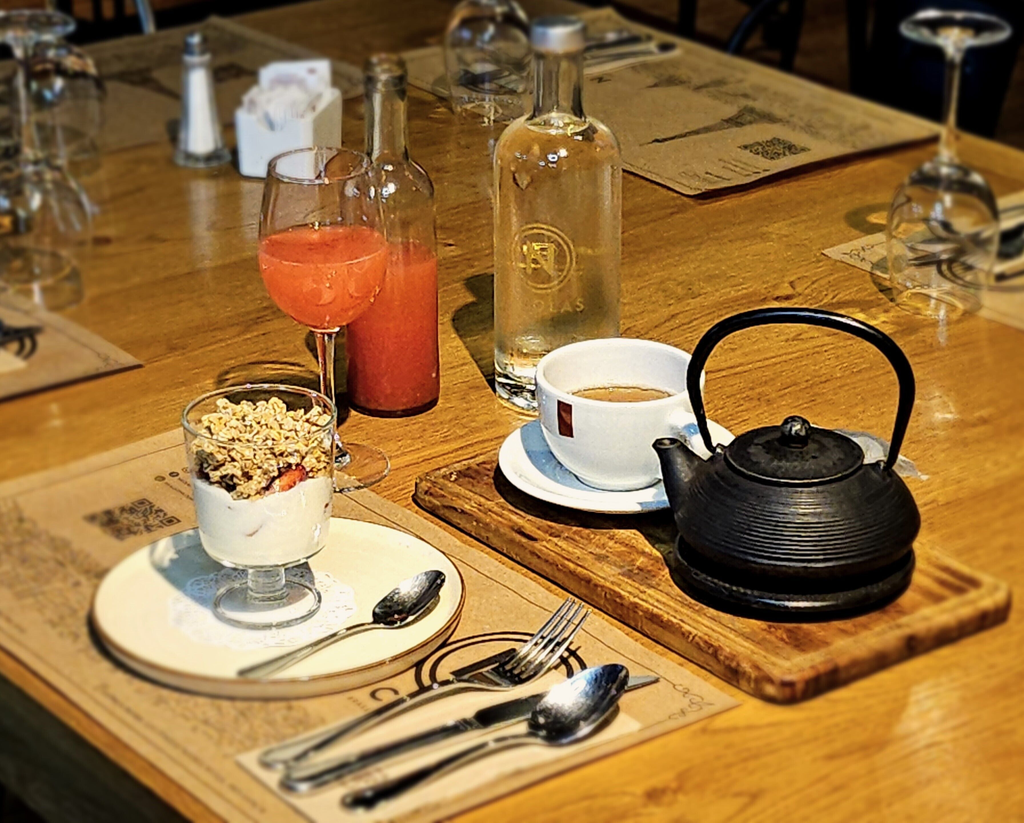 A wooden table with a teapot, a cup of coffee, a glass of juice, and a dessert with a spoon.