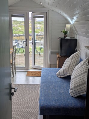 A room with a bed, a television on a stand, and a view through a door to an outdoor area with chairs and a table. - Pod Beag Beside the Sea, Isle of Eriskay (Eriskay)