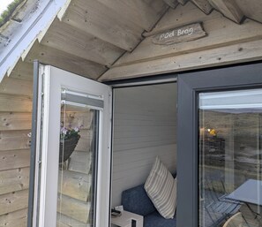 A wooden exterior with a sliding glass door, a bench with a striped cushion, and a sign above the door. - Pod Beag Beside the Sea, Isle of Eriskay (Eriskay)