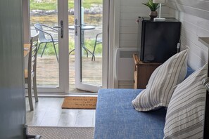 A room with a blue sofa, a television on a wooden stand, a small table with a plant, and a door leading to an outdoor area with chairs and a table. - Pod Beag Beside the Sea, Isle of Eriskay (Eriskay)