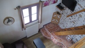 A room with a wooden floor, a brown sofa, a small table, a window with curtains, a radiator, a television on the wall, and a staircase with a wooden handrail. - Camping La Chevauchée (Bussière-Dunoise)