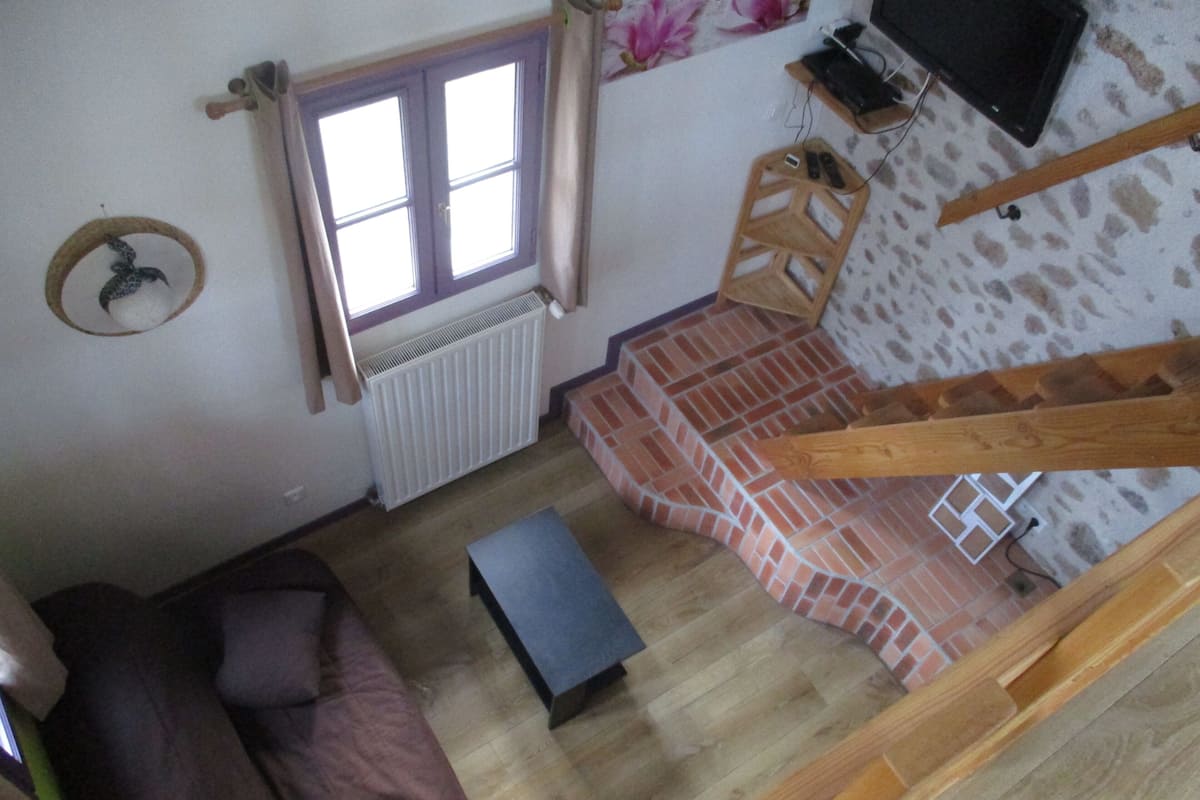 A room with a wooden floor, a brown sofa, a small table, a window with curtains, a radiator, a television on the wall, and a staircase with a wooden handrail.