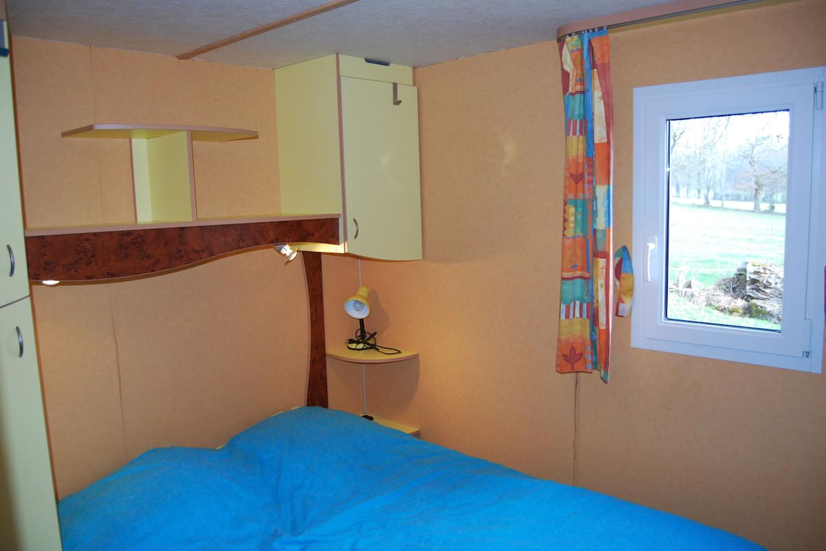 A bedroom with a bed, a desk with a lamp, a window with a view of trees, and a door.