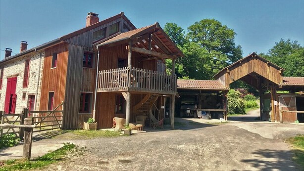 A rustic wooden building with a balcony, a water wheel, and a covered area with a red door. - Camping La Chevauchée (Bussière-Dunoise)