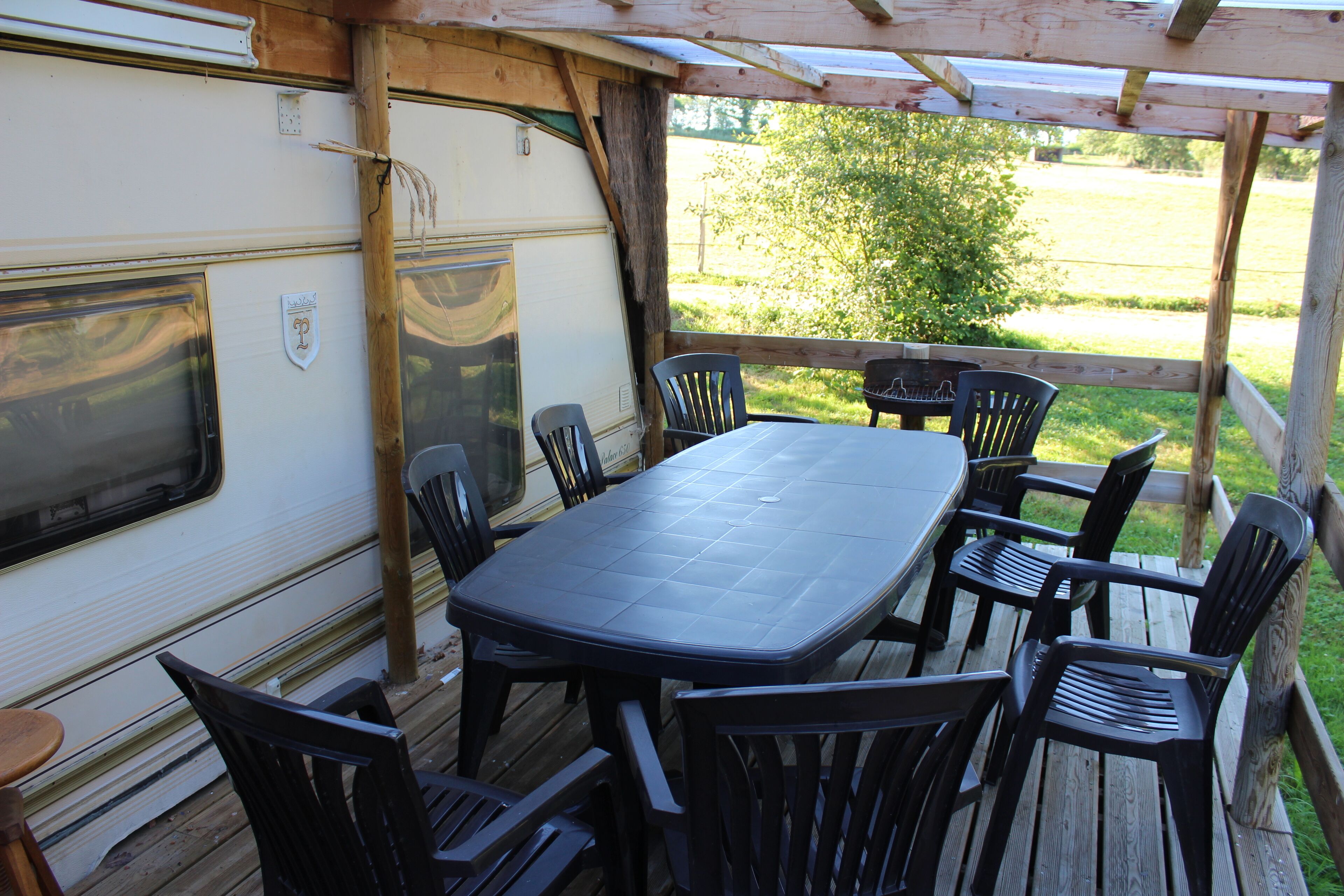 Een overdekt terras met een tafel en stoelen, een houten terras en een witte camper.