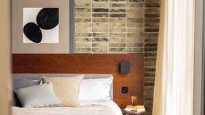 A hotel room with a brick-patterned wall, a wooden headboard, a bed with multiple pillows, a bedside table with a book and a glass, and a window with curtains. - The Hamlet (Geneva)