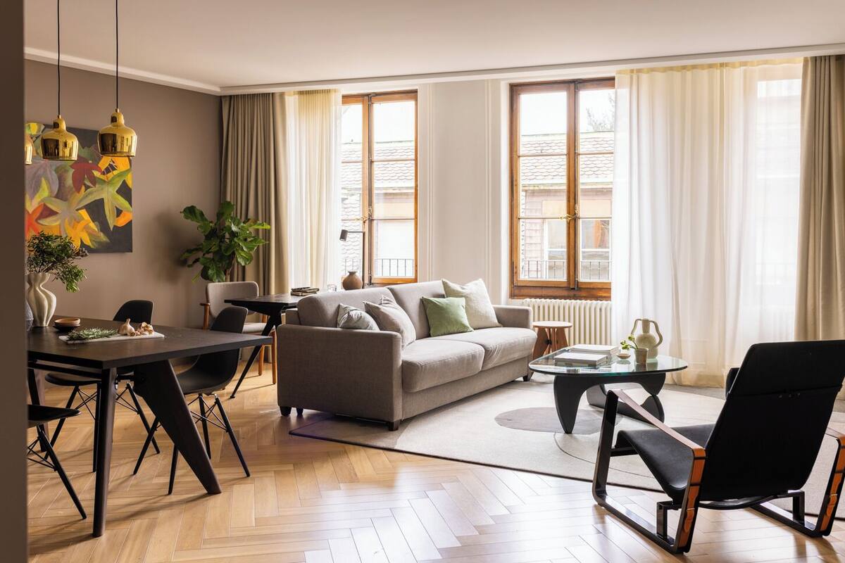 A modern living room with a sofa, coffee table, dining table, and chairs. There are large windows with curtains, a plant, and artwork on the wall.