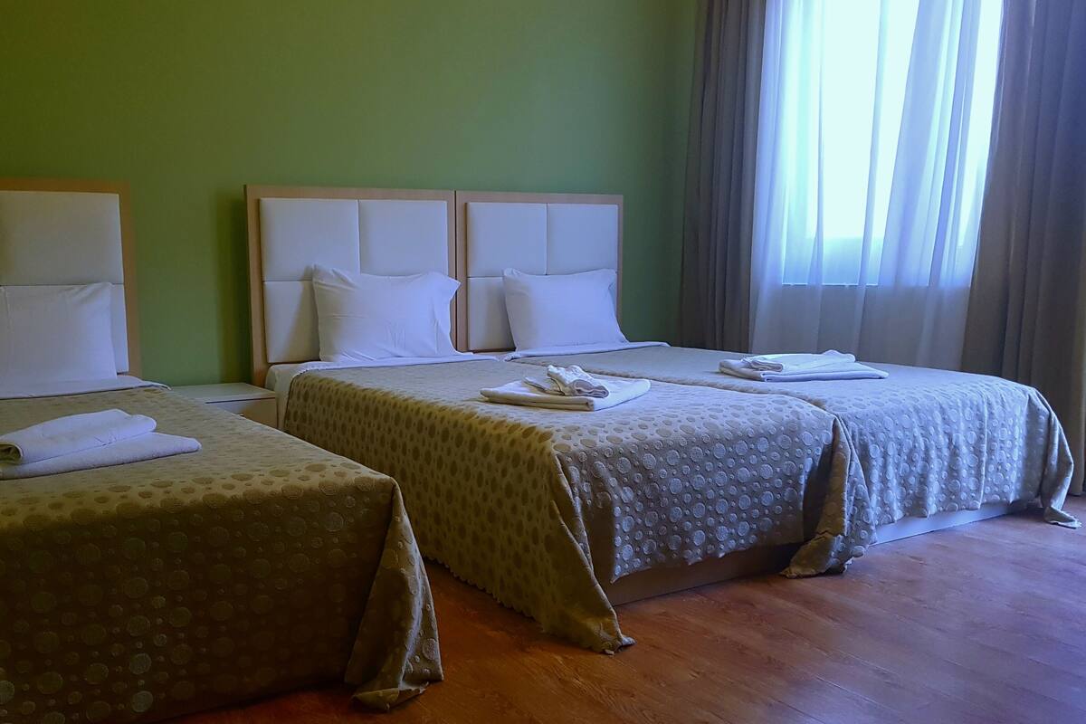 A hotel room with two single beds, each with a headboard, white bedding, and folded towels. The room has a wooden floor and green walls.