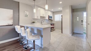 A modern kitchen with white cabinetry, a central island with bar stools, pendant lighting, and a view into the adjacent living area.
