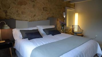 A neatly made bed with white and grey bedding, a stone wall, a bedside lamp, and a chair with a vase of flowers.