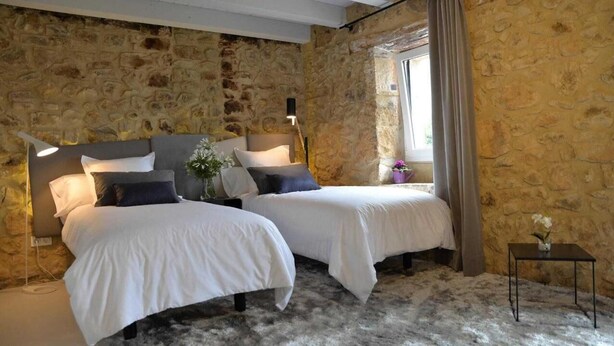 A room with two beds, stone walls, a small table with a vase, and a window with curtains. - Hotel Garaiko Landetxea (Garai)