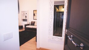 A hallway with a dark wooden door, a white wall with a decorative mirror, and a black rug leading to a living area with a sofa and a bookshelf. - Ludovisi Decò (Rome)