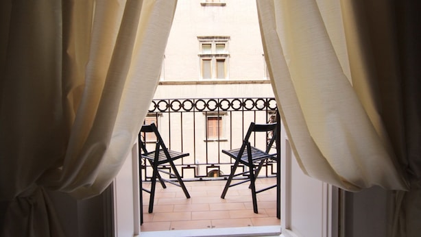 Uitzicht door een raam met gordijnen, waarop een balkon met twee stoelen en een gebouw op de achtergrond te zien is.