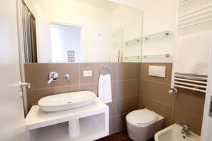 Ein modernes Badezimmer mit einem weißen Waschbecken, einer Toilette und einem Bidet. Über dem Waschbecken befinden sich Glastrensen und an der Wand ein Handtuchhalter.