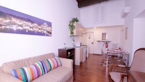 A living room with a beige sofa, a colorful striped pillow, a dining area with a table and chairs, a kitchen with a microwave, and a hanging plant. - Monticelli Viola (Rome)