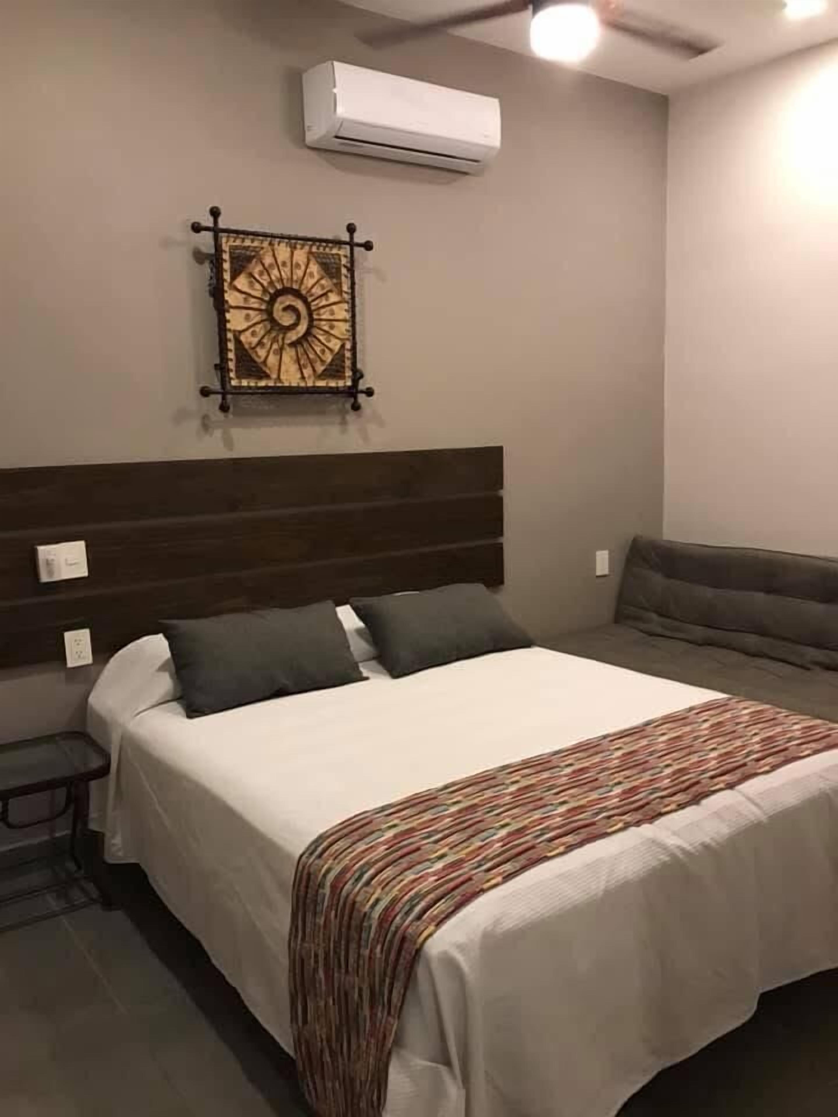 Habitación de hotel con una cama grande, cabecera, una mesita, un banco y un equipo de aire acondicionado en la pared.