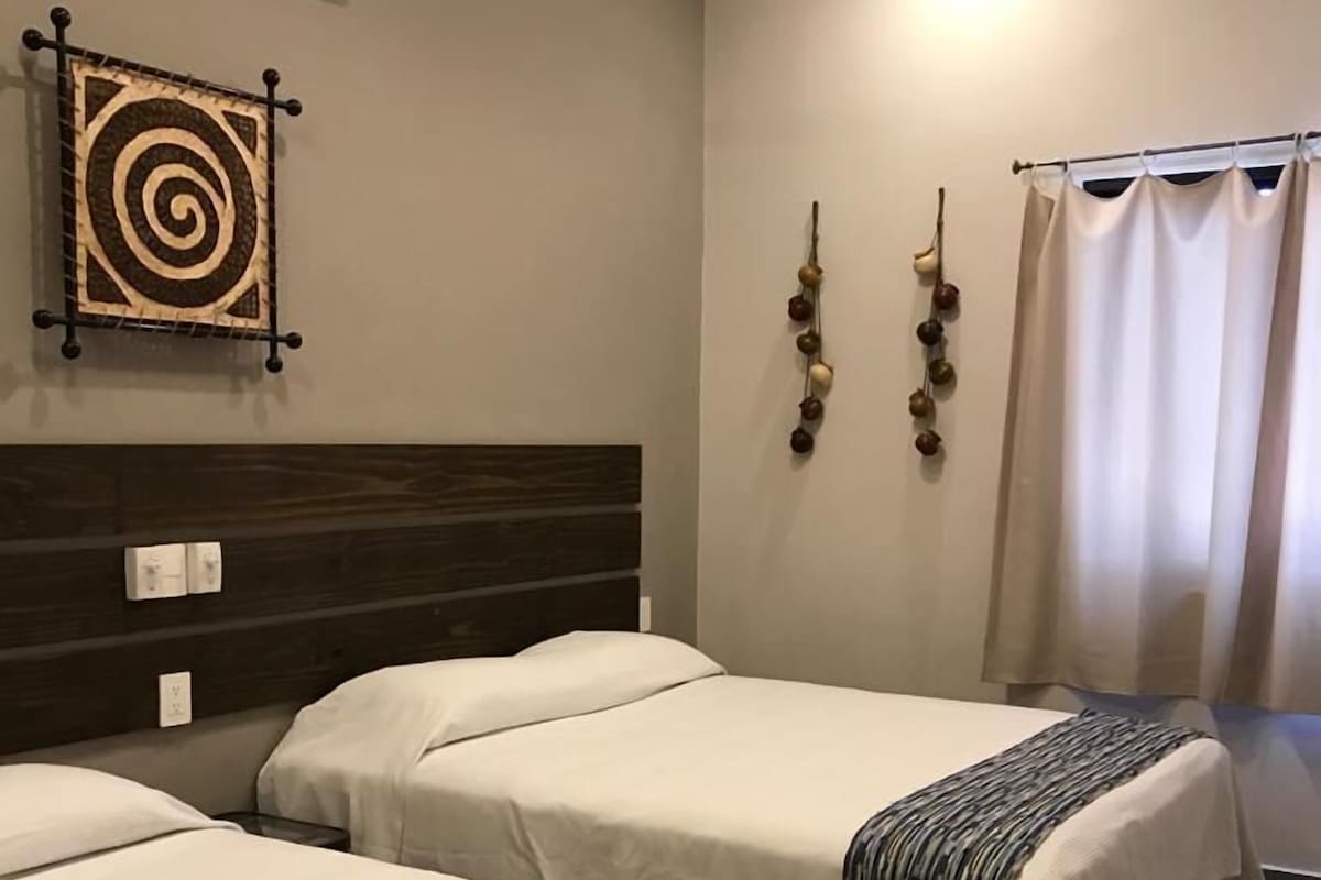 Habitación de hotel con dos camas, ventilador de techo, aire acondicionado, un tapiz decorativo y una cortina.