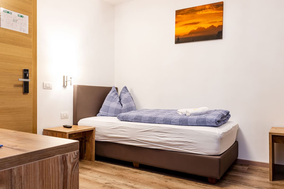 Una cama individual con cabecera, una mesita de noche, un escritorio de madera y un cuadro de un atardecer en la pared.