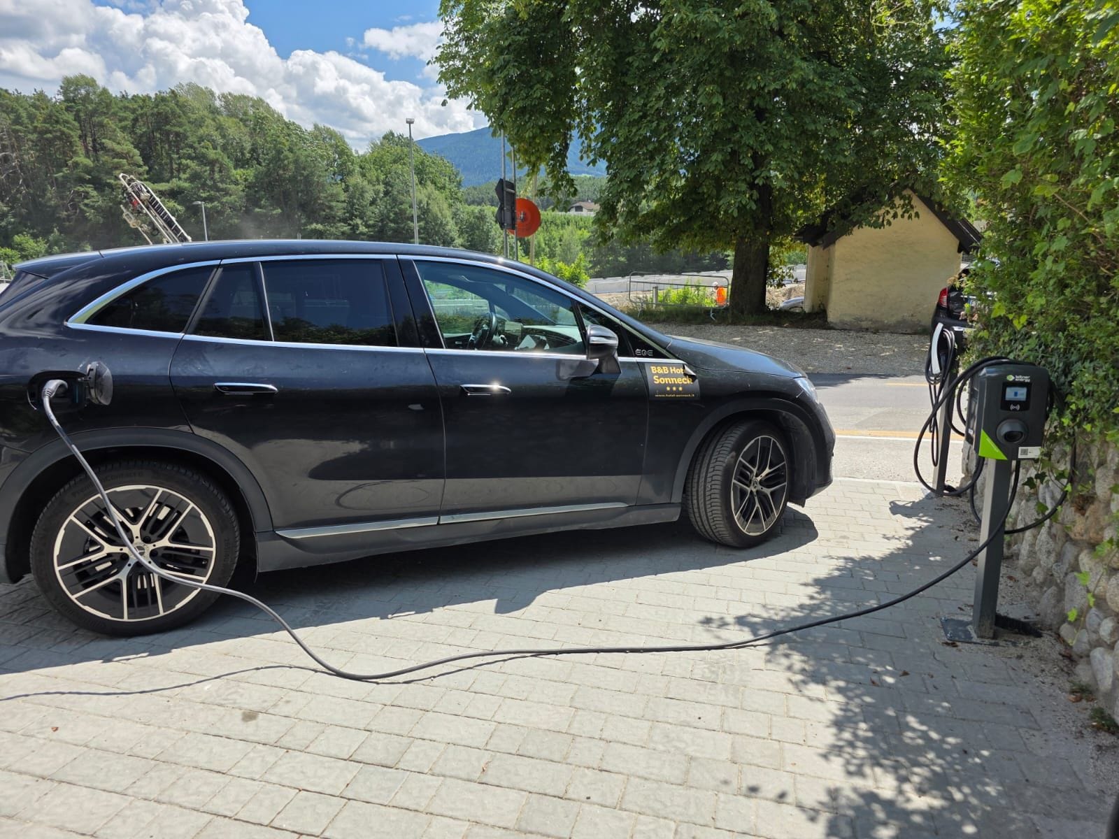 Un coche negro estacionado en una estación de carga para vehículos eléctricos, con pavimento de ladrillo y árboles al fondo.