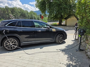 Une voiture noire est garée à une borne de recharge pour véhicules électriques, sur un trottoir pavé de briques, avec des arbres en arrière-plan.