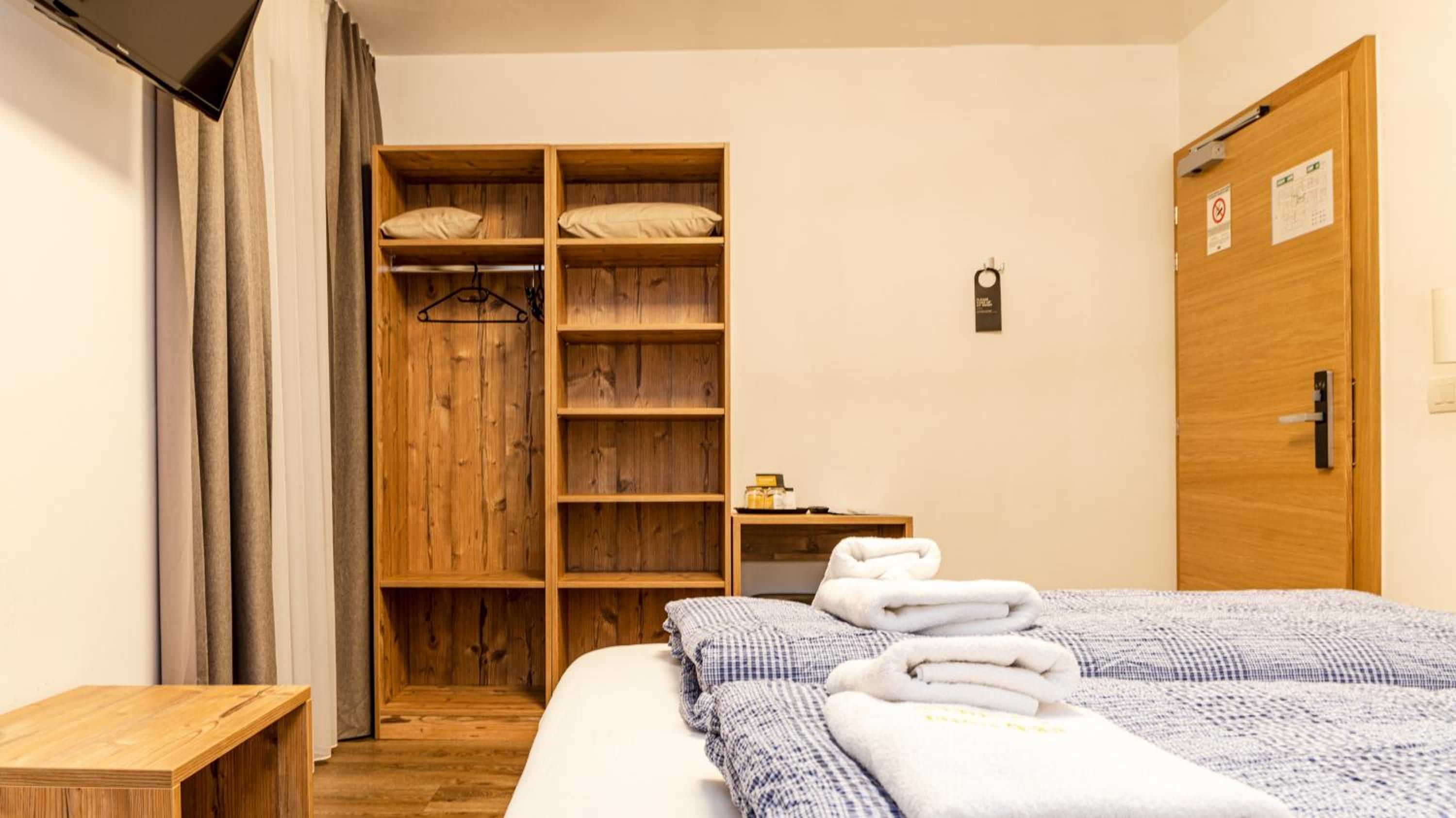 A hotel room with a wooden wardrobe, a bed with towels, a bedside table, and a door with a sign.