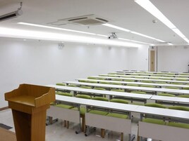 Uma sala de conferências com mesas e cadeiras dispostas em fileiras, um púlpito e uma porta ao fundo.