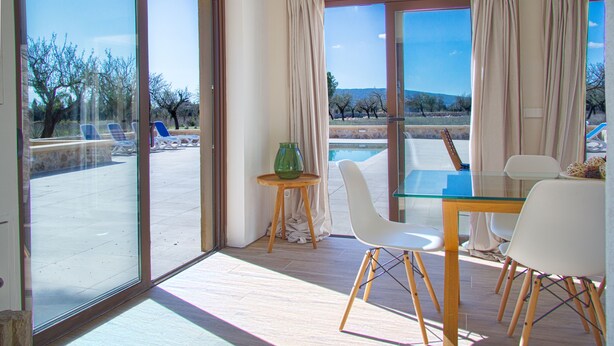 A dining area with a glass table and chairs, a small side table with a vase, and a view of the outdoor patio with chairs and a pool. - Finca Sa Caseta Biniali (Sencelles)