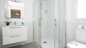 A modern bathroom with a glass shower enclosure, white tiles, a white vanity with drawers, a potted plant, and a toilet with a blue sticker. - MONKÓ KIWI TROPYCAL APARTMENT (Torremolinos)