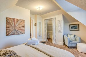 A bedroom with a bed, a chair, a painting, and a door leading to another room.
