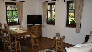 Een woonkamer met een houten eettafel, stoelen, een flatscreen televisie, een bank en twee ramen met gordijnen.