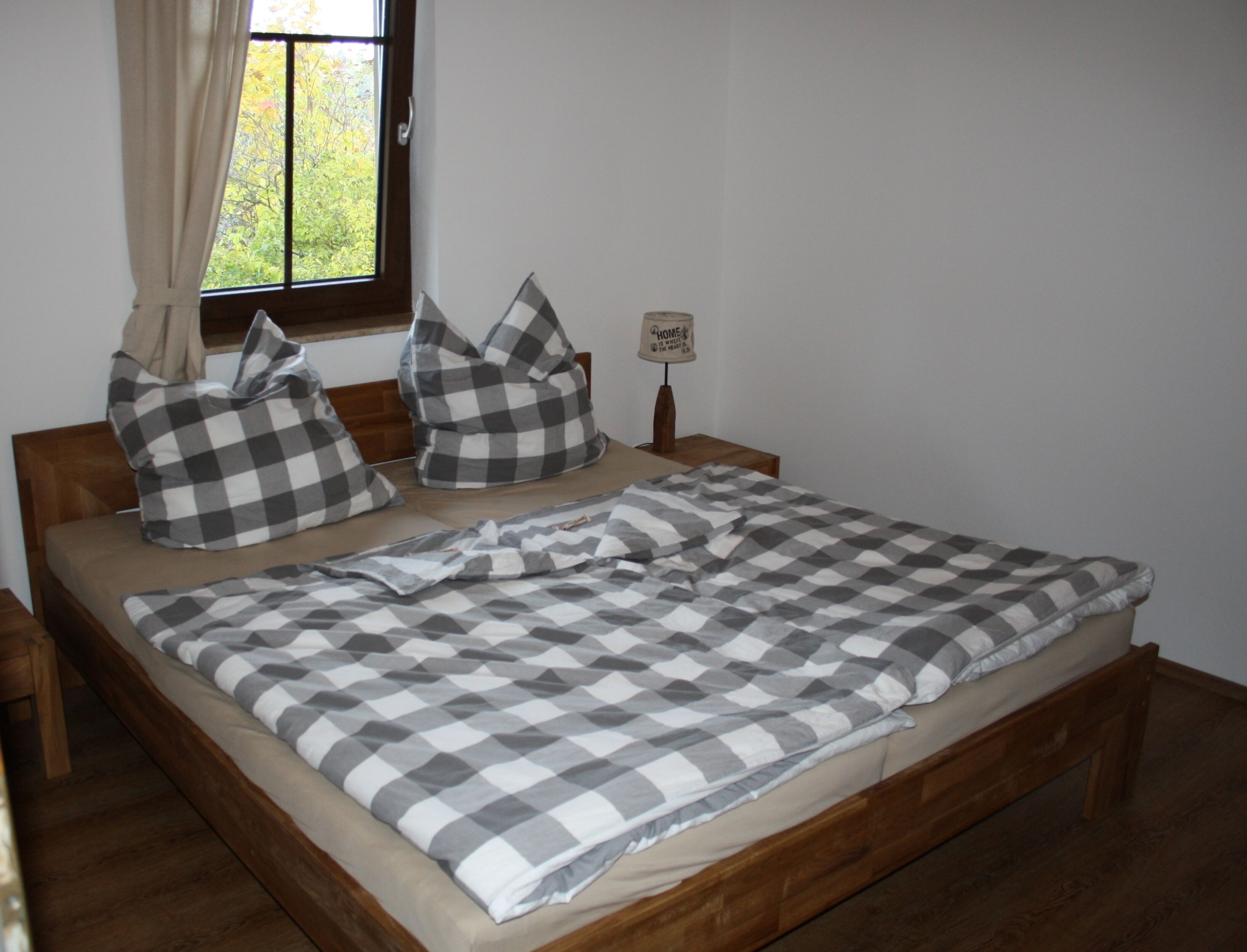 A neatly made bed with a checkered bedspread and pillows, a wooden headboard, a bedside table with a lamp, and a window with curtains.