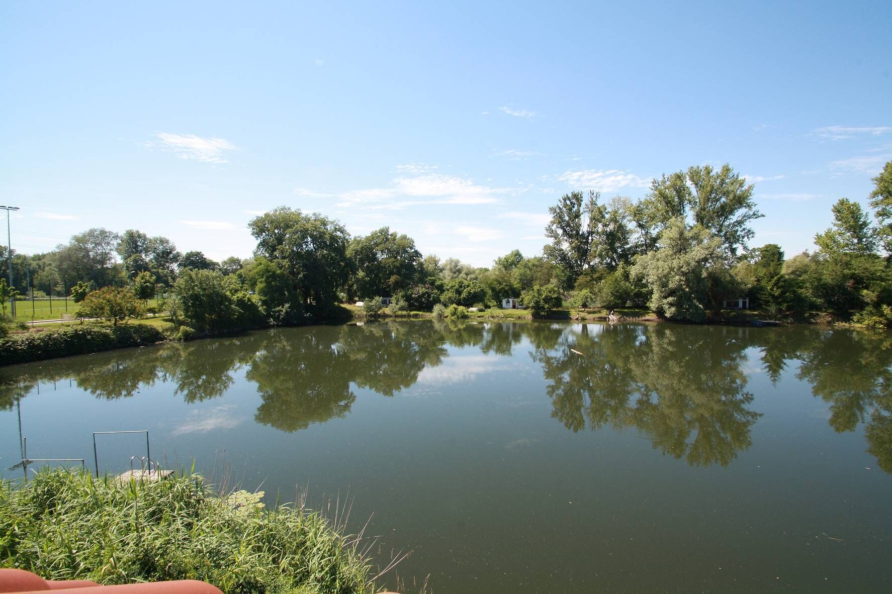 Un lac paisible, dont les arbres se reflètent dans l’eau, entouré de verdure et surplombé d’un ciel dégagé.