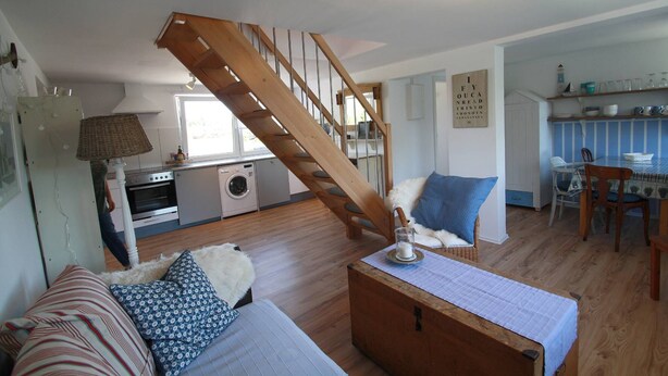 A cozy living room with a wooden staircase, a sofa with patterned cushions, a dining table with chairs, and a kitchen area with a washing machine and oven. - Maisonettewohnung am Privatsee (Karlsruhe)