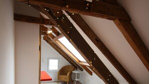 A bedroom with a sloped ceiling, wooden beams, a bed with a patterned blanket, a chair, a small table, and a window with natural light. - Maisonettewohnung mit großer Terrasse (Karlsruhe)
