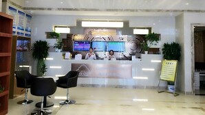 Zona de recepción de hotel con diseño moderno, que incluye un mostrador, dos monitores y una pantalla digital. Se observan dos recepcionistas, una estantería de madera y dos sillas negras en primer plano.